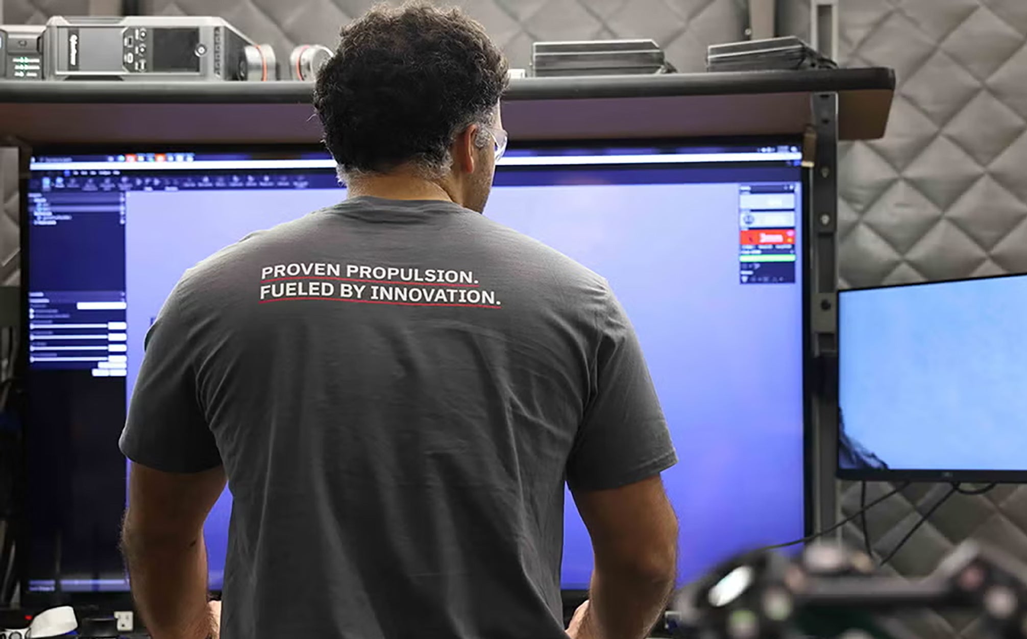 An employee looks at his screen in a T-shirt that says "Proven Propulsion. Fueled by Innovation."