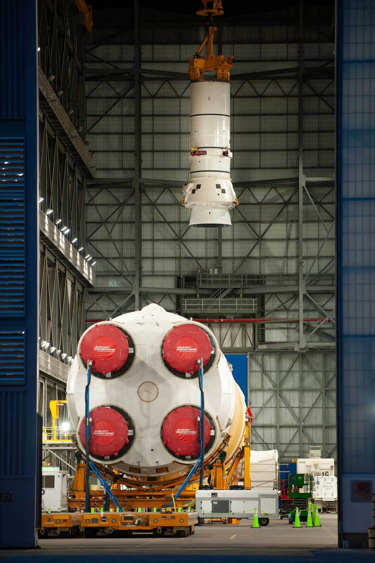  Photo Credit: NASA. Down the transfer aisle from the Artemis II SLS core stage, an overhead crane hoists the left aft assembly, or bottom portion of the solid rocket boosters for the SLS moon rocket  inside the Vehicle Assembly Building at NASA’s Kennedy Space Center.
