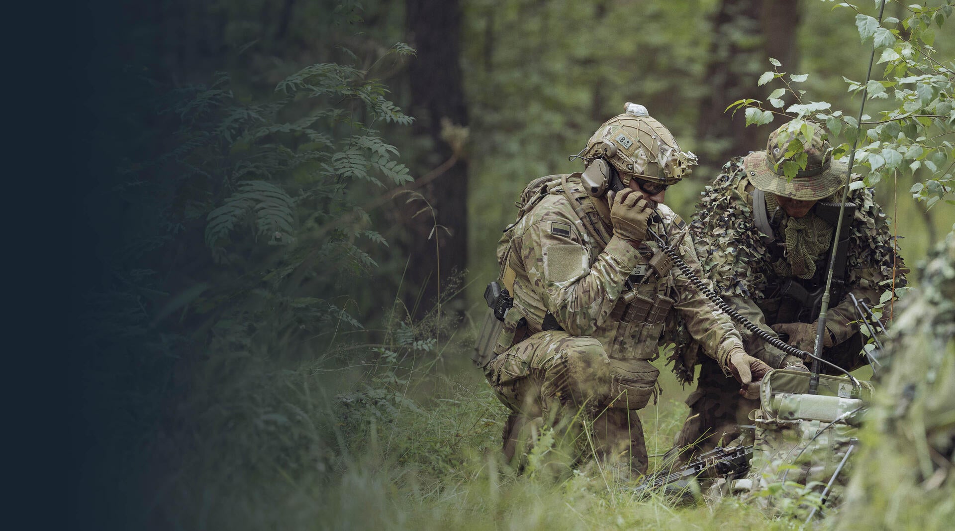 Two soldiers speak on tactical radios in a forest
