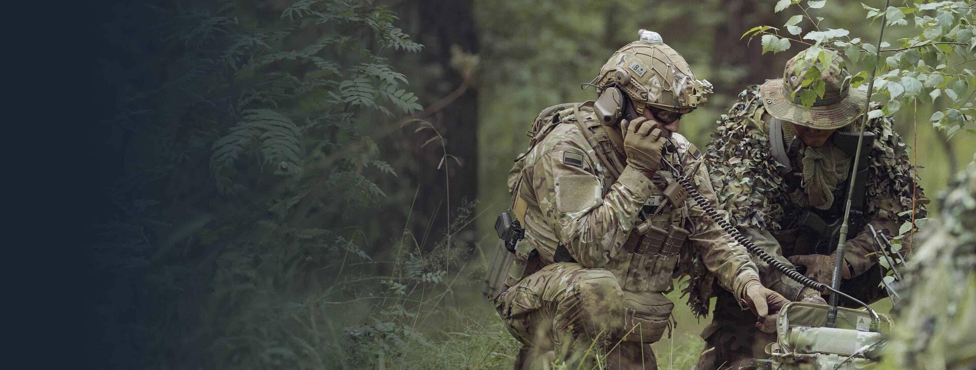Two soldiers speak on tactical radios in a forest