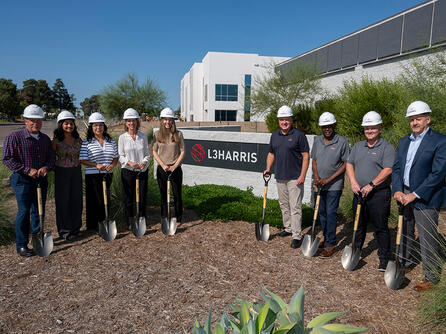 Employees stand in front of an L3Harris sign with shovels and hard hats