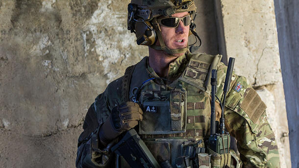 Soldier in front of concrete wall with camouflaged gear and L3Harris tactical radio displayed